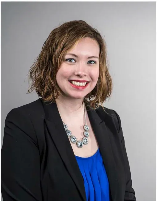 Smiling woman in a blue blouse and dark blazer against a plain gray background