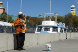 A musician in a colorful shirt and hat plays a saxophone by the waterfront, setting the perfect backdrop for event photography. With a boat and tower silhouetted in the background, his donation bucket sits beneath him on this bright, sunny day.