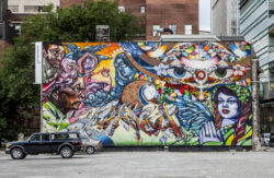 A vibrant mural on a brick wall depicts surreal faces, abstract designs, and colorful patterns. An eye-catching blend of blues, reds, and oranges fills the wall, resembling event photographys vivid storytelling. A parked black vehicle is in front, with urban buildings in the background.