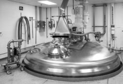 A black and white image captures a large, shiny stainless steel industrial mixer in a factory setting, highlighting the expertise akin to corporate photography services. The mixer boasts various pipes and controls with a smooth, domed lid. The room features white tiled walls and visible wiring and equipment.
