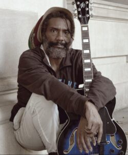 A person with a beard and dreadlocks is sitting on the ground, leaning against a stone wall. They are wearing a hat, dark jacket, and light pants, holding a blue electric guitar upright beside them—capturing an unplugged moment akin to candid event photography.