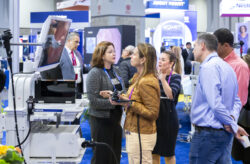 A group of people stands around a medical equipment display at a trade show, captured perfectly in event photography. They are engaged in a discussion while one person holds a control device. The background features various exhibition booths and signs, adding depth to the conference photography setting.