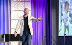 A person in a checkered blazer and jeans stands on stage, gesturing with their right hand. Behind them is a large screen displaying an image of a child wearing a shirt that says super loved. Captured with precision in this event photography setting, the scene is framed by elegant purple curtains.